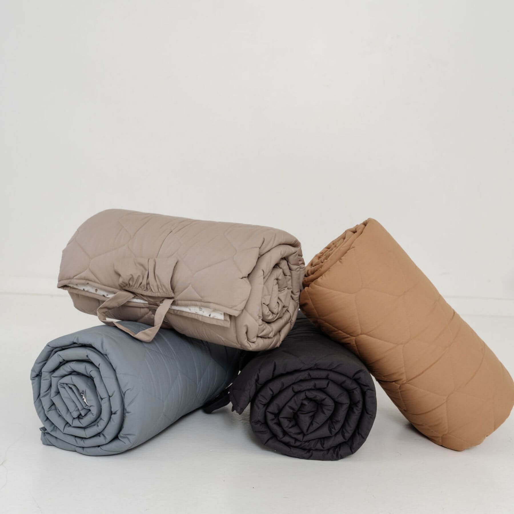 Four rolled-up blankets in brown, gray, and black on a light gray background