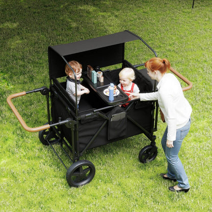 ToyRides Elite Wagon Stroller with 4-Seater - Black is shown outdoors on grass, with two children seated inside and a woman standing beside it holding the tray.
