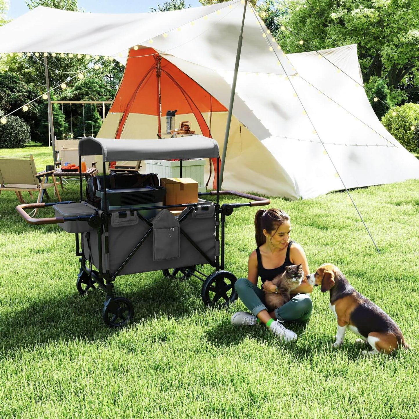 A woman sits on a green lawn with a cat and a dog beside ToyRides Elite Wagon Stroller with 4-Seater - Gray, with a white and orange camping tent and a large white canopy in the background.