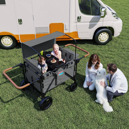 ToyRides Elite Wagon Stroller with 4-Seater - Gray is shown on a grassy lawn in front of a white and orange RV. Two children are sitting inside the wagon, while a woman and man kneel beside it petting a white dog.