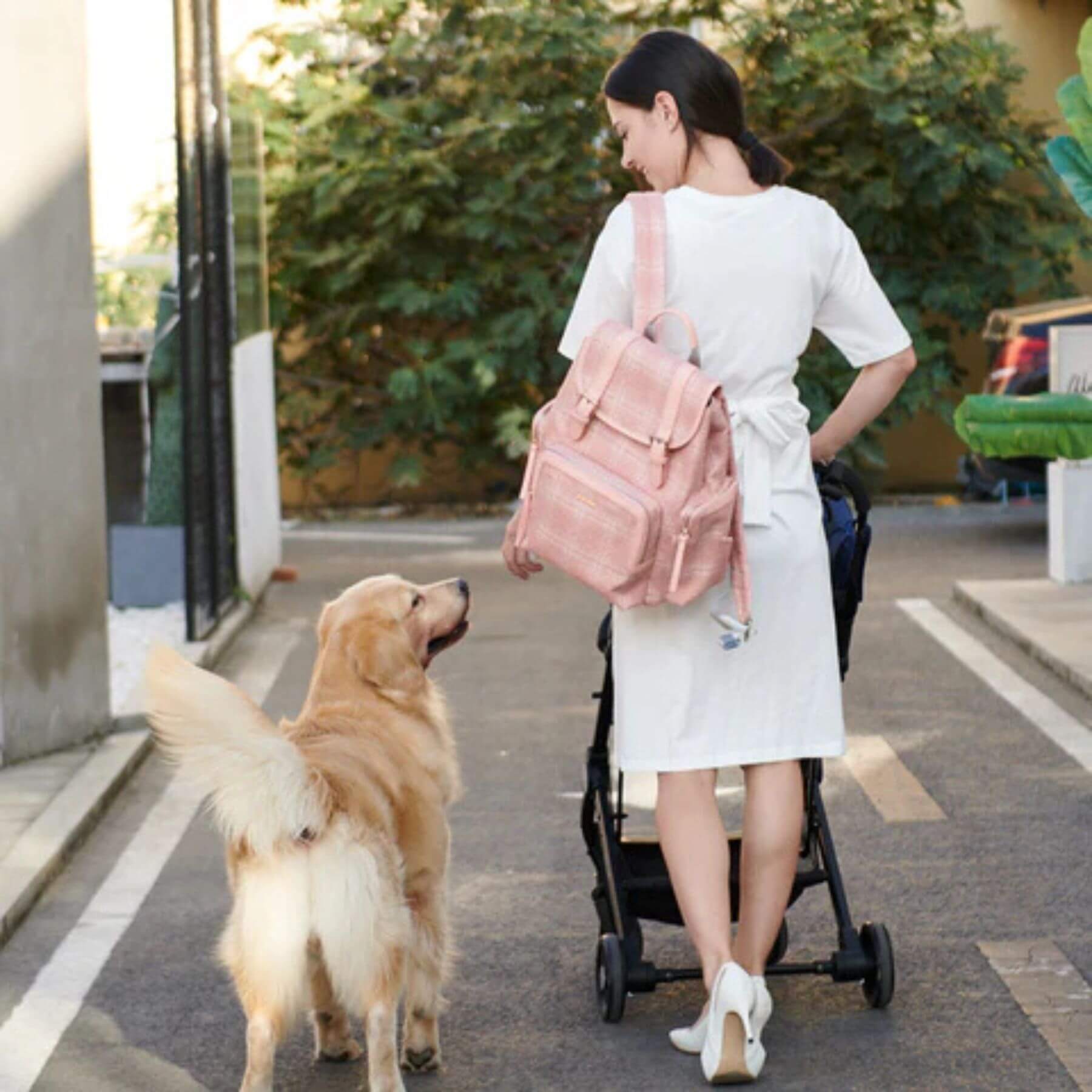 Woman with Sunveno Tweed Diaper Backpack Pink and stroller walking a dog on a sidewalk.