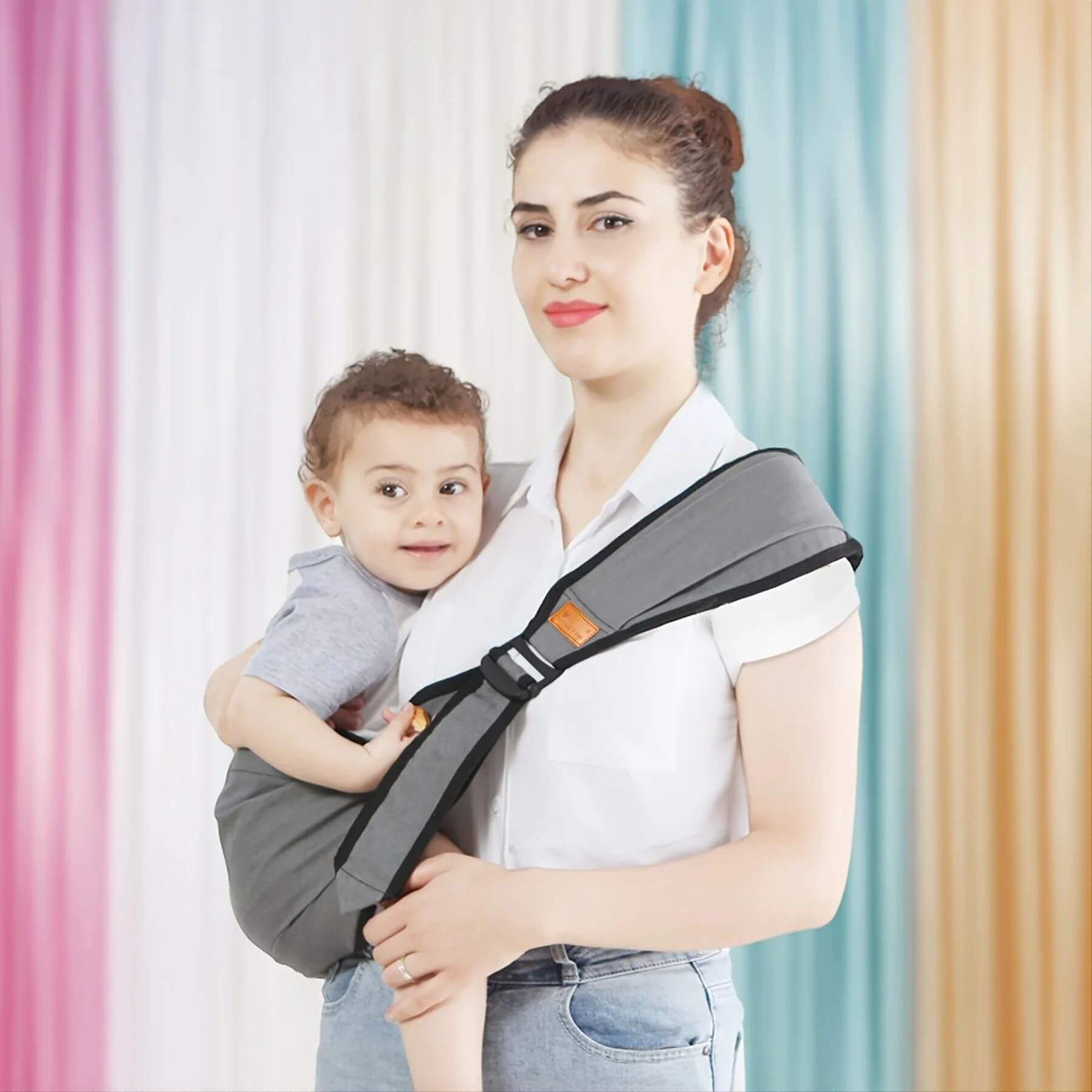 Woman holding a child using RocketBaby USA Universal Baby Carrier Grey against a colorful curtain background