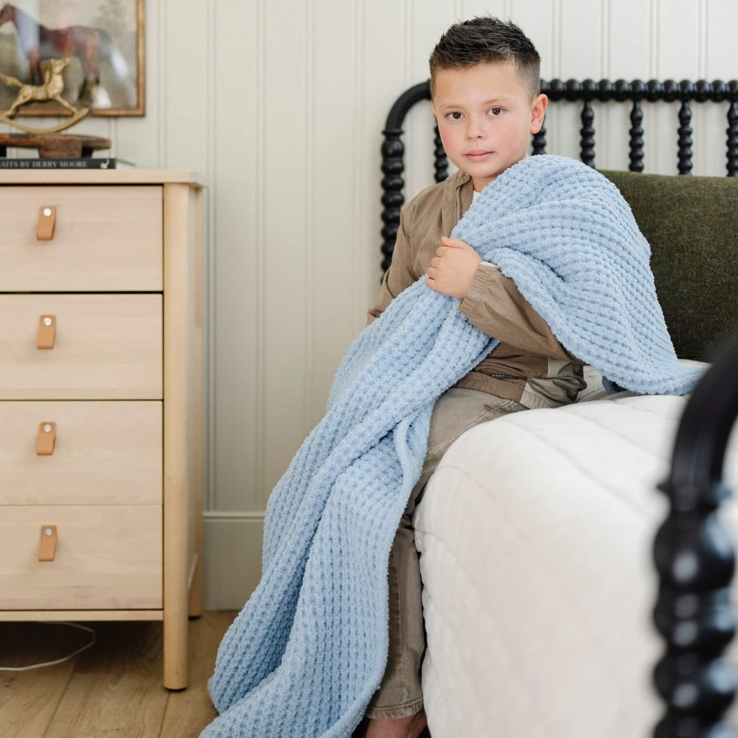 Child sitting on a couch wrapped in Saranoni Waffle Knit Toddler Blanket - Blue Jay in a cozy living room.