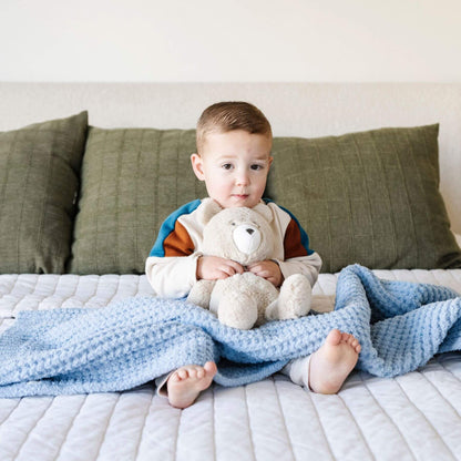 Child sitting on a bed holding a teddy bear with Saranoni Waffle Knit Toddler Blanket - Blue Jay