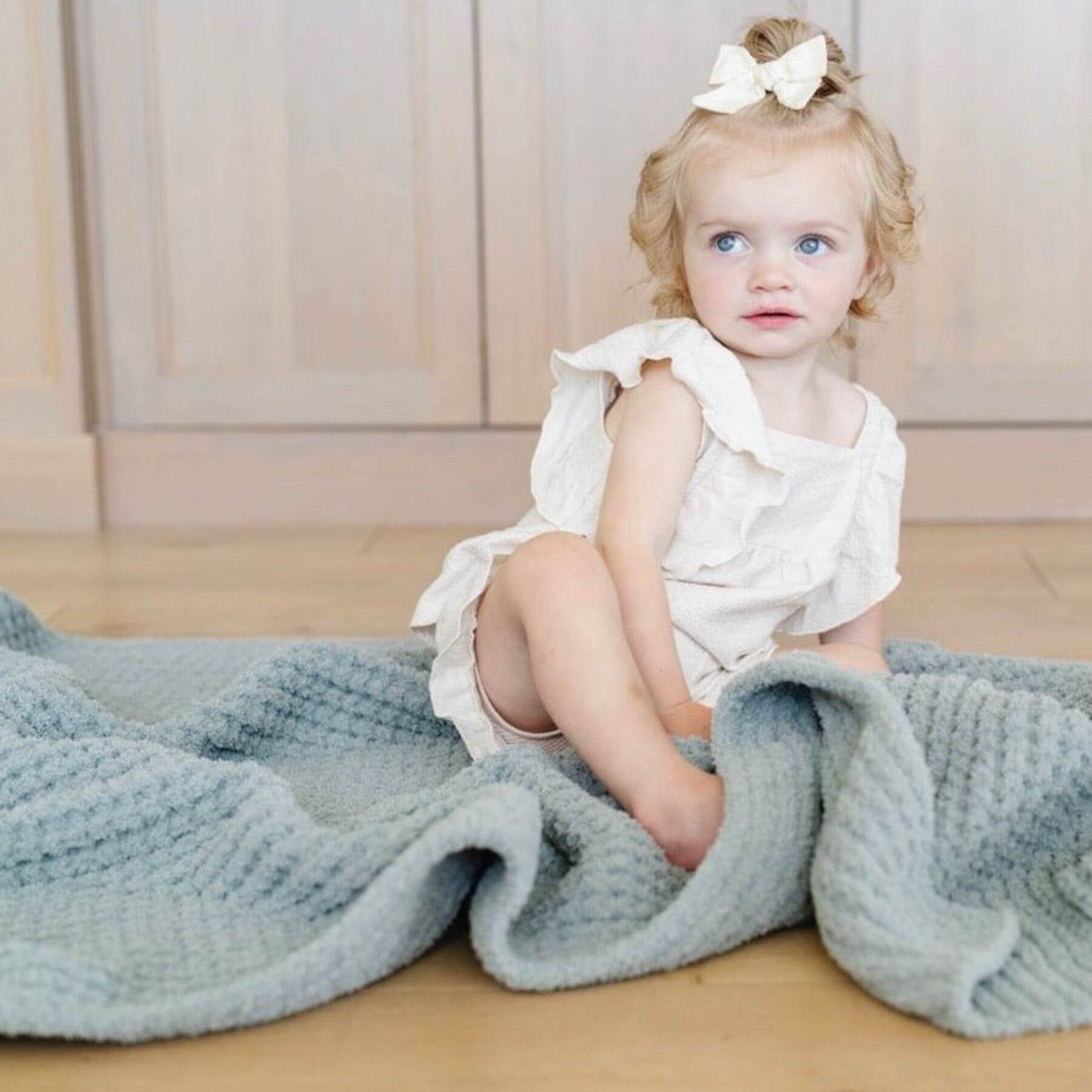 Child in a white outfit sitting on Saranoni Waffle Knit Toddler Blanket - Elm indoors.