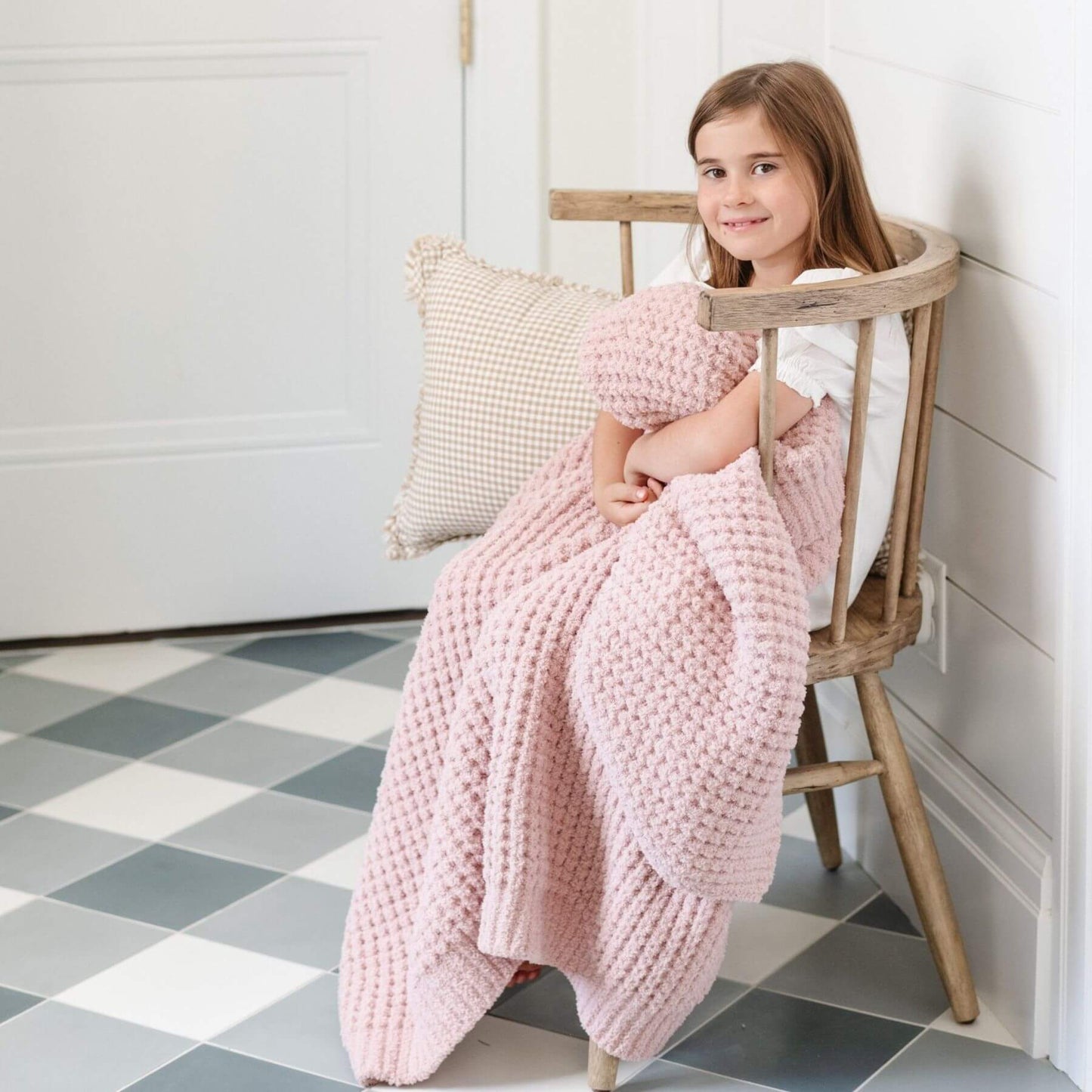 Young girl sitting on a wooden chair with Saranoni Waffle Knit Toddler Blanket - Rosette and pillow in a room with checkered floor.