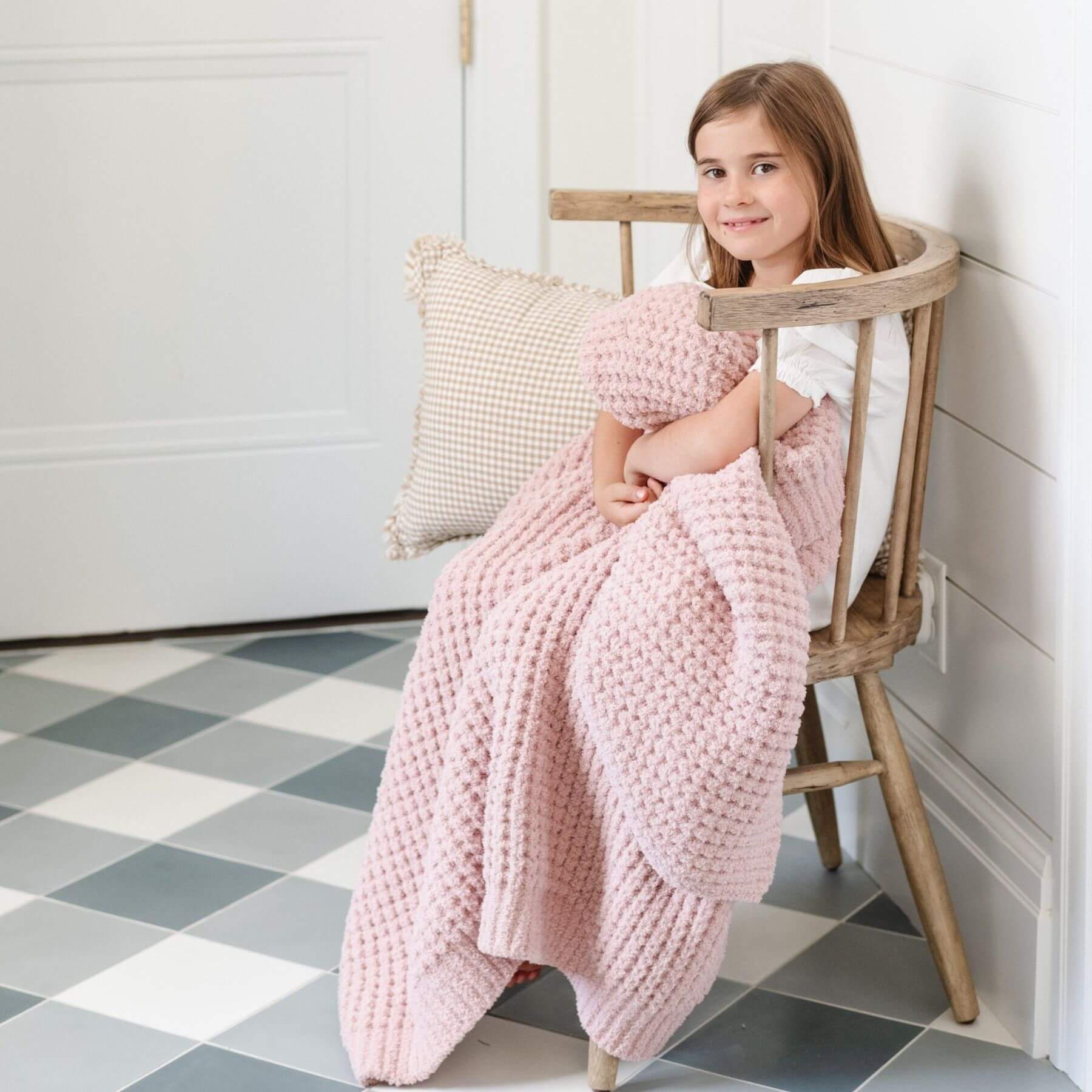 Young girl sitting on a wooden chair with Saranoni Waffle Knit Toddler Blanket - Rosette and pillow in a room with checkered floor.
