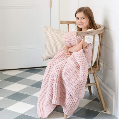 Young girl sitting on a wooden chair with Saranoni Waffle Knit Toddler Blanket - Rosette and pillow in a room with checkered floor.