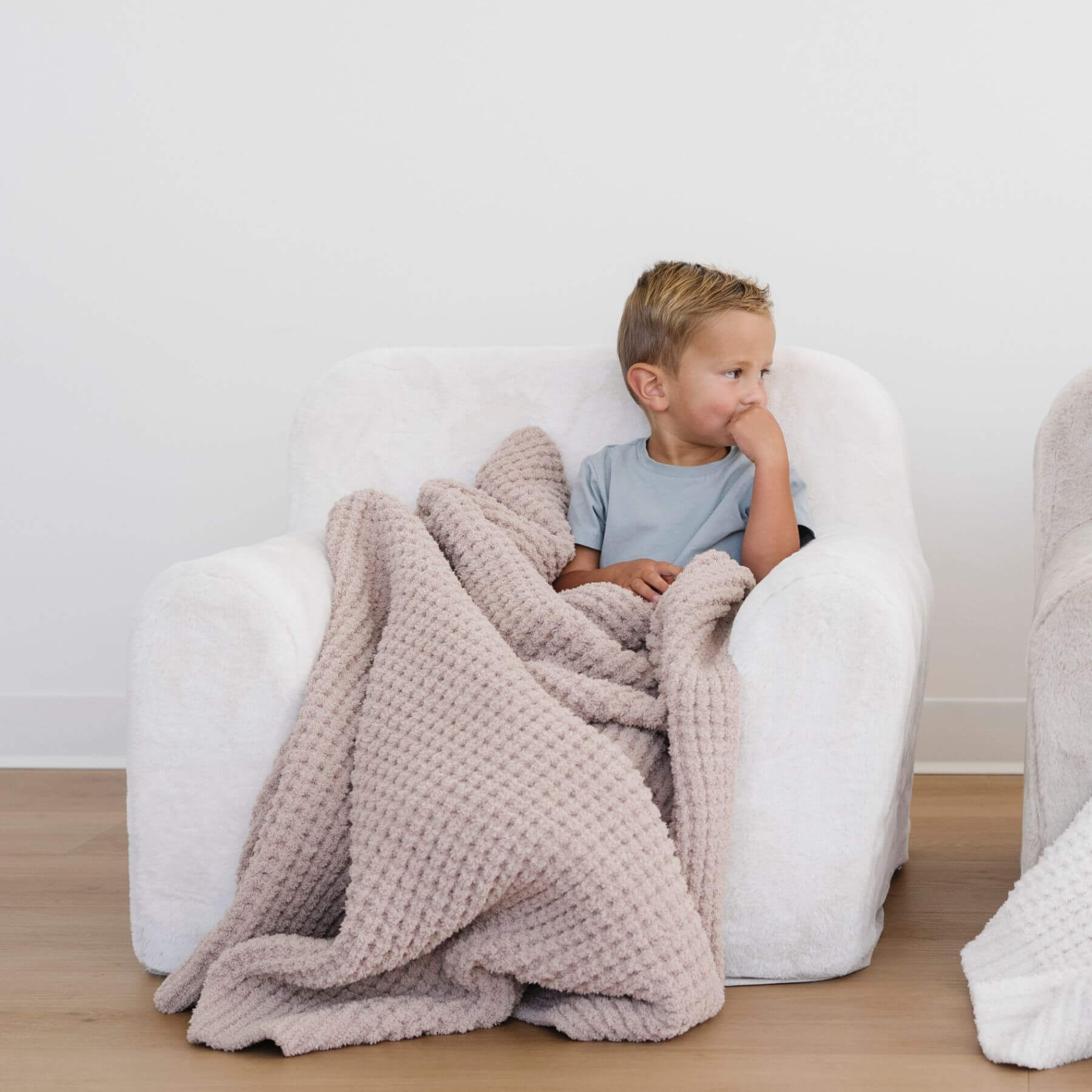 Child sitting on a white chair with Saranoni Waffle Knit Toddler Blanket - Wheat, in a minimalistic room.