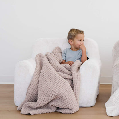 Child sitting on a white chair with Saranoni Waffle Knit Toddler Blanket - Wheat, in a minimalistic room.