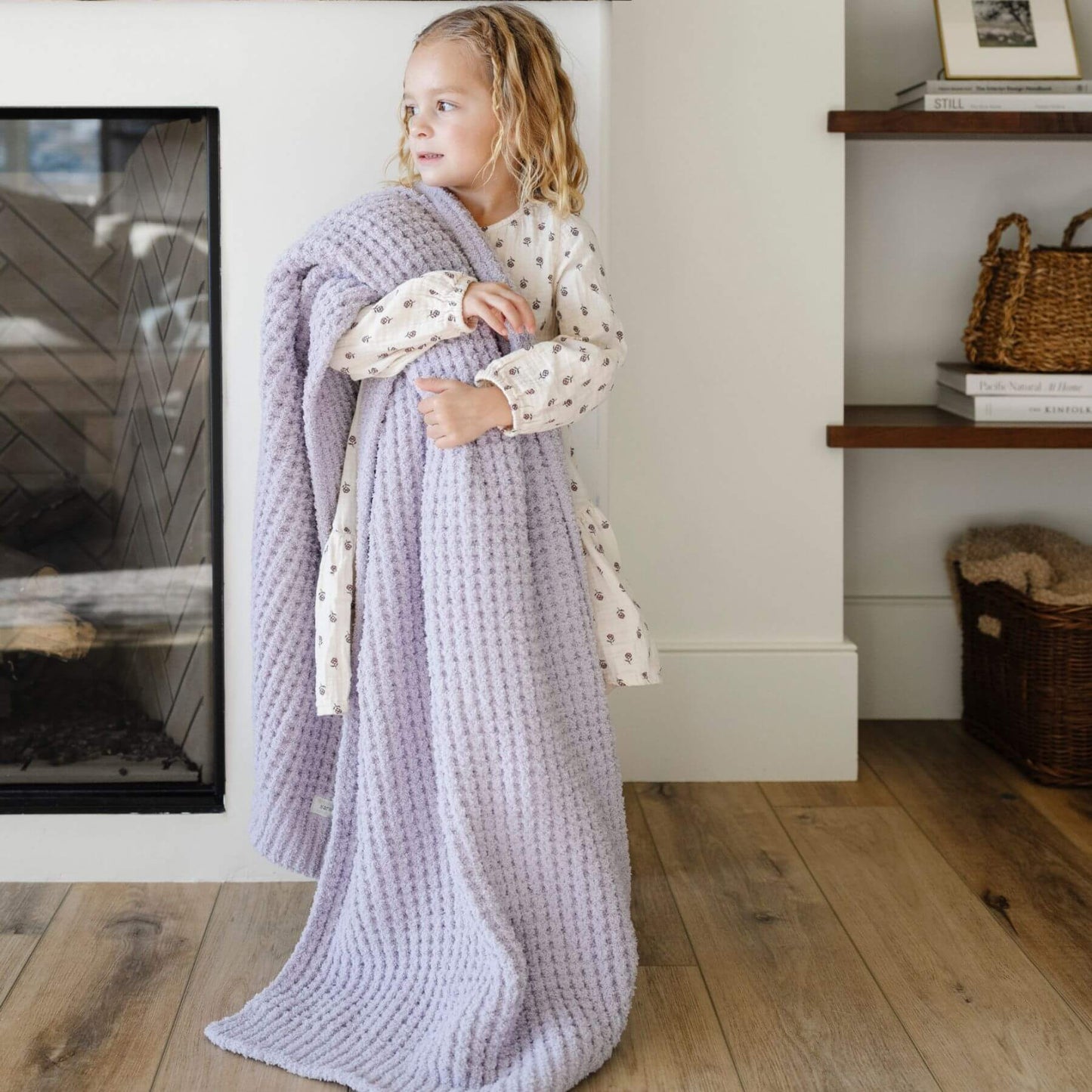Child wrapped in Saranoni Waffle Knit Toddler Blanket - Wisteria standing in a cozy room with a fireplace and shelves.