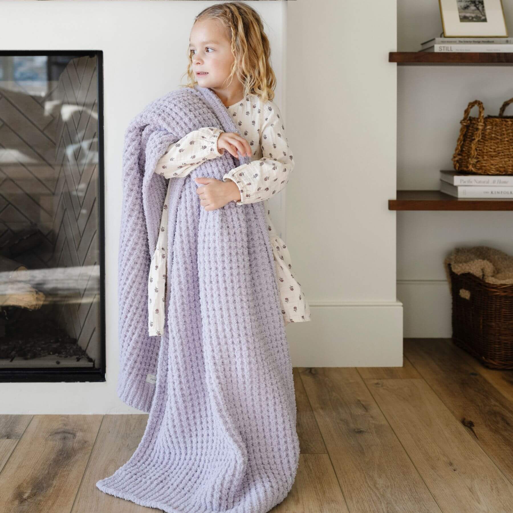 Child wrapped in Saranoni Waffle Knit Toddler Blanket - Wisteria standing in a cozy room with a fireplace and shelves.