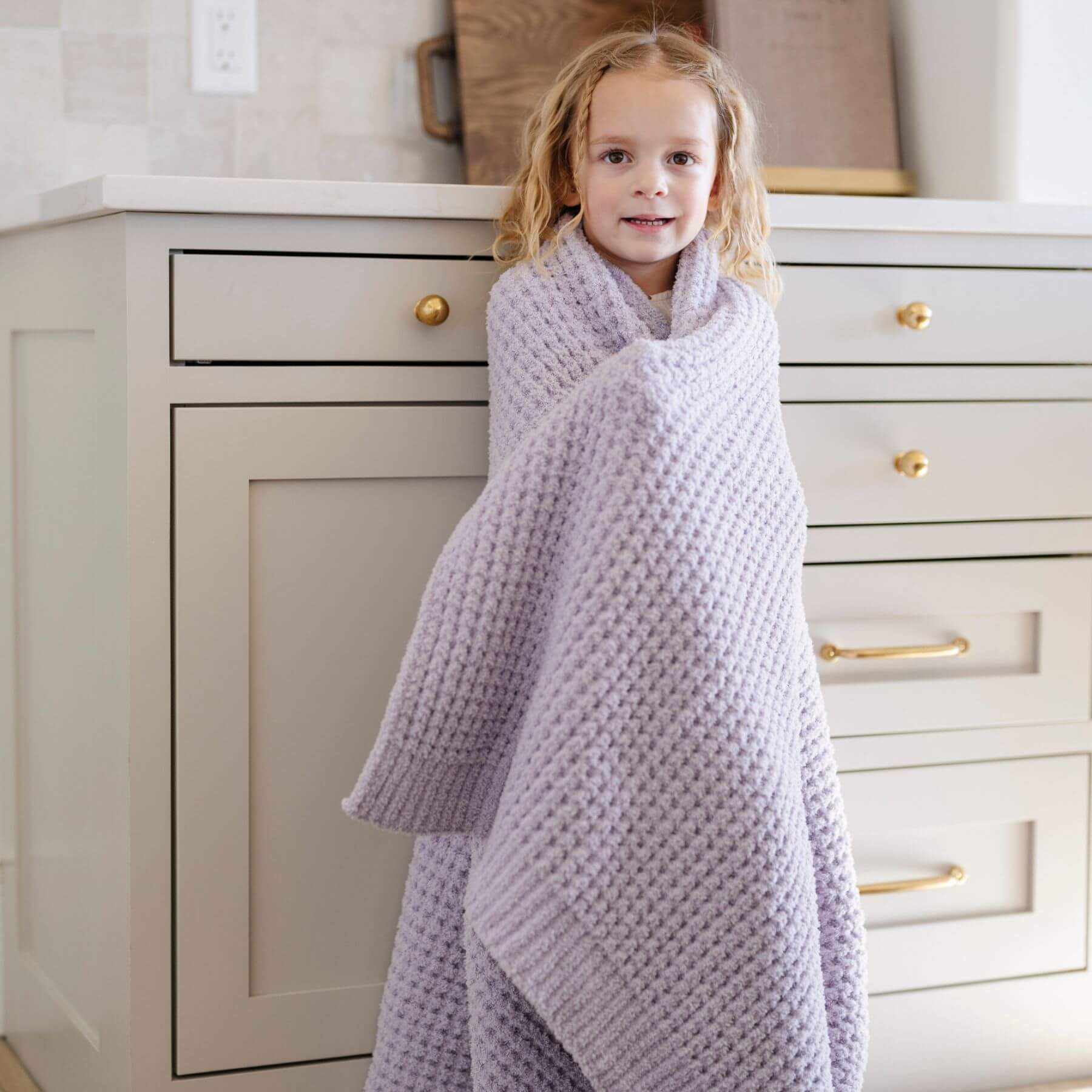Child wrapped in Saranoni Waffle Knit Toddler Blanket - Wisteria standing in a room with white cabinets.