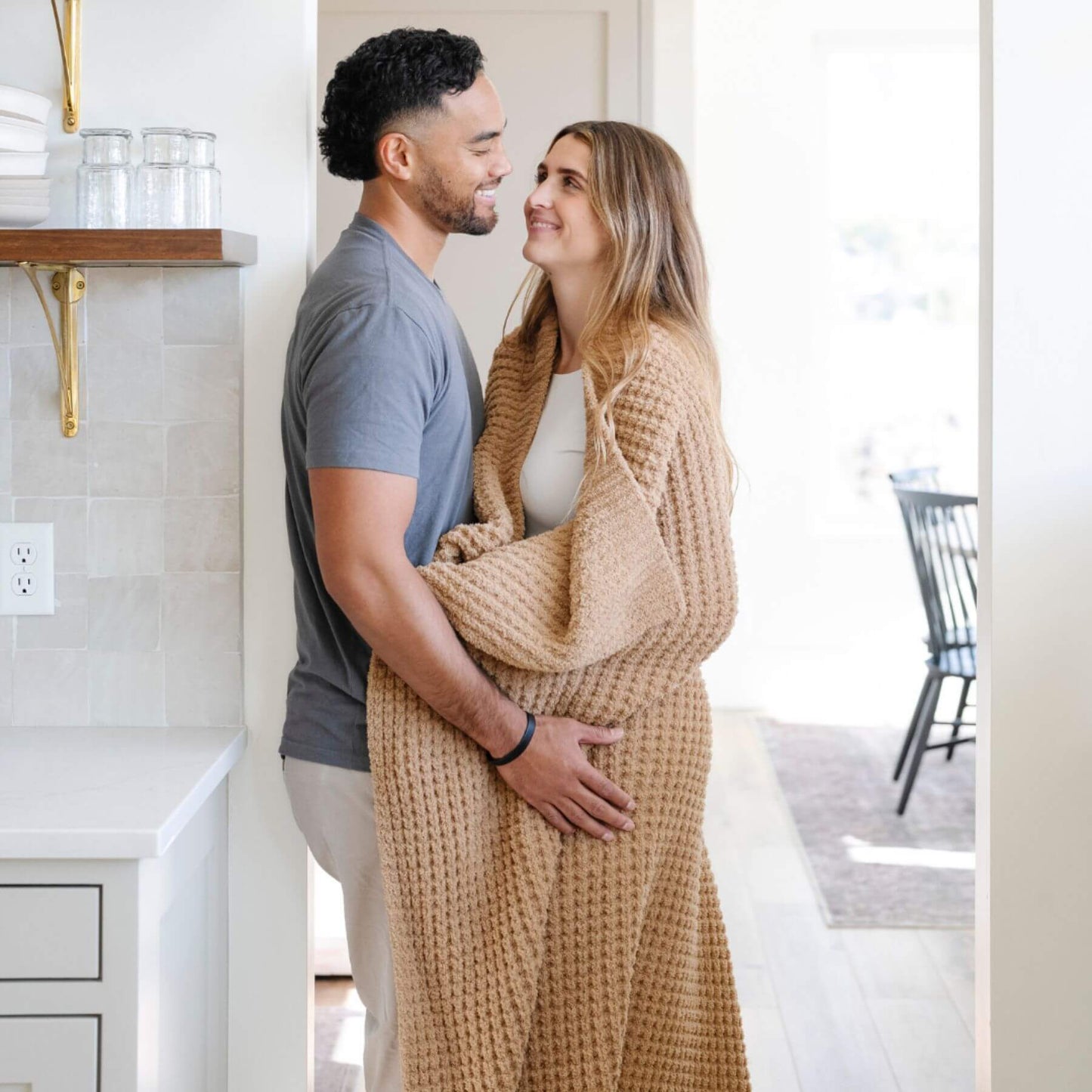 Man and woman standing close together in a kitchen, with the woman wrapped in Saranoni Waffle Knit XL Throw Blanket - Allspice
