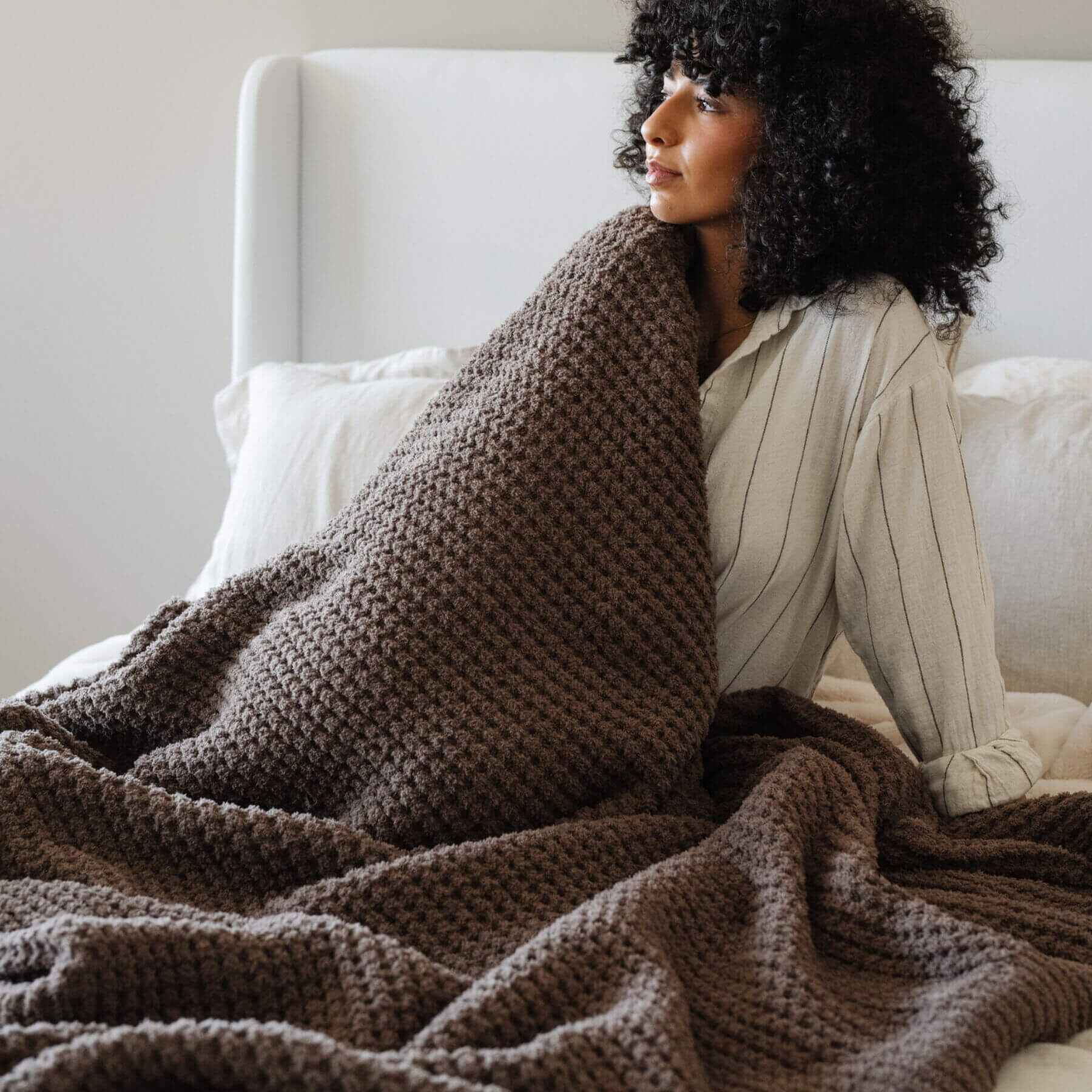 Woman wrapped in Saranoni Waffle Knit XL Throw Blanket - Chestnut sitting on a couch.