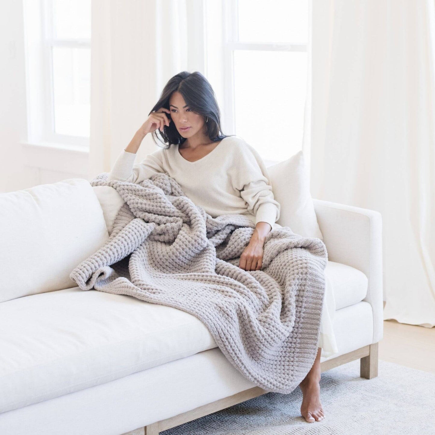 Woman sitting on a white couch with Saranoni Waffle Knit XL Throw Blanket - Moonbeam draped over her, in a bright room.