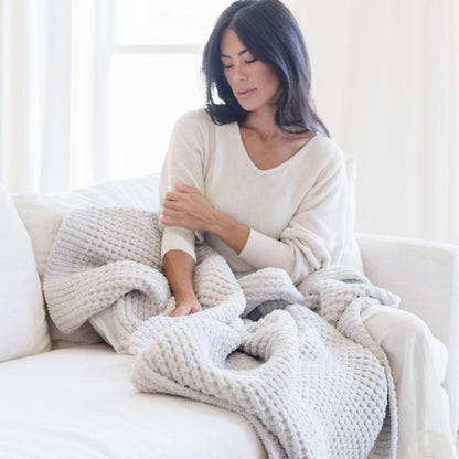 Woman sitting on a couch wrapped in Saranoni Waffle Knit XL Throw Blanket - Moonbeam in a bright room