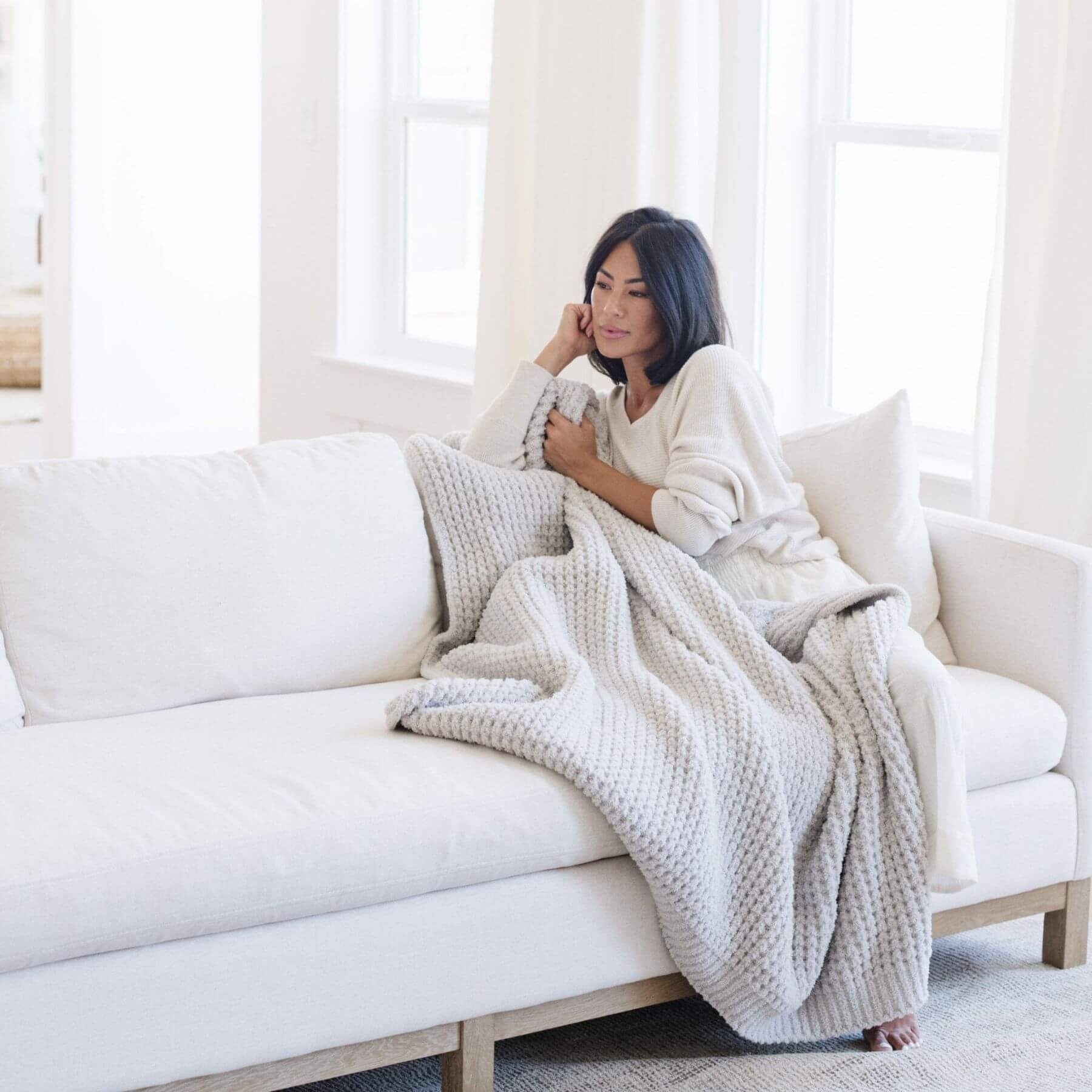 Woman sitting on a white couch with Saranoni Waffle Knit XL Throw Blanket - Moonbeam in a bright room