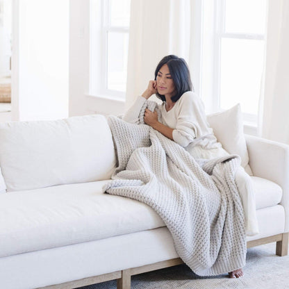 Woman sitting on a white couch with Saranoni Waffle Knit XL Throw Blanket - Moonbeam in a bright room