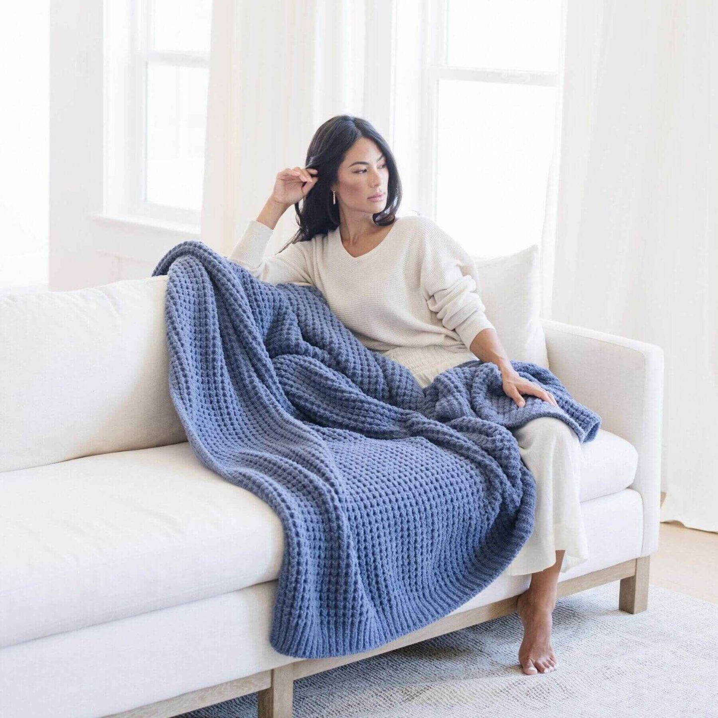Woman sitting on a white couch with Saranoni Waffle Knit XL Throw Blanket - Oxford draped over her, in a bright room.