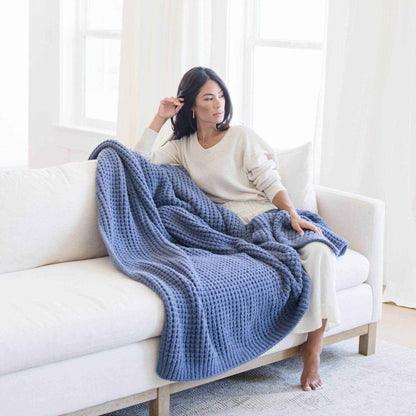 Woman sitting on a white couch with Saranoni Waffle Knit XL Throw Blanket - Oxford draped over her, in a bright room.