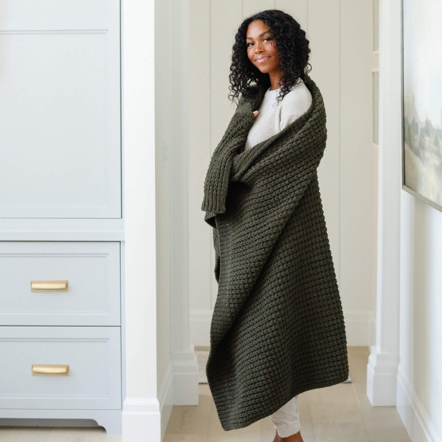 Woman wearing Saranoni Waffle Knit XL Throw Blanket - Thyme in a room with light gray walls and white trim.