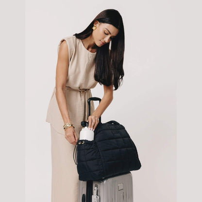 Woman holding augustnoa the weekender duffle bag black over a suitcase on a plain background