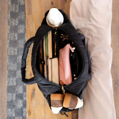 augustnoa the weekender duffle bag black with various items including a pink wallet and a white helmet on a wooden floor.