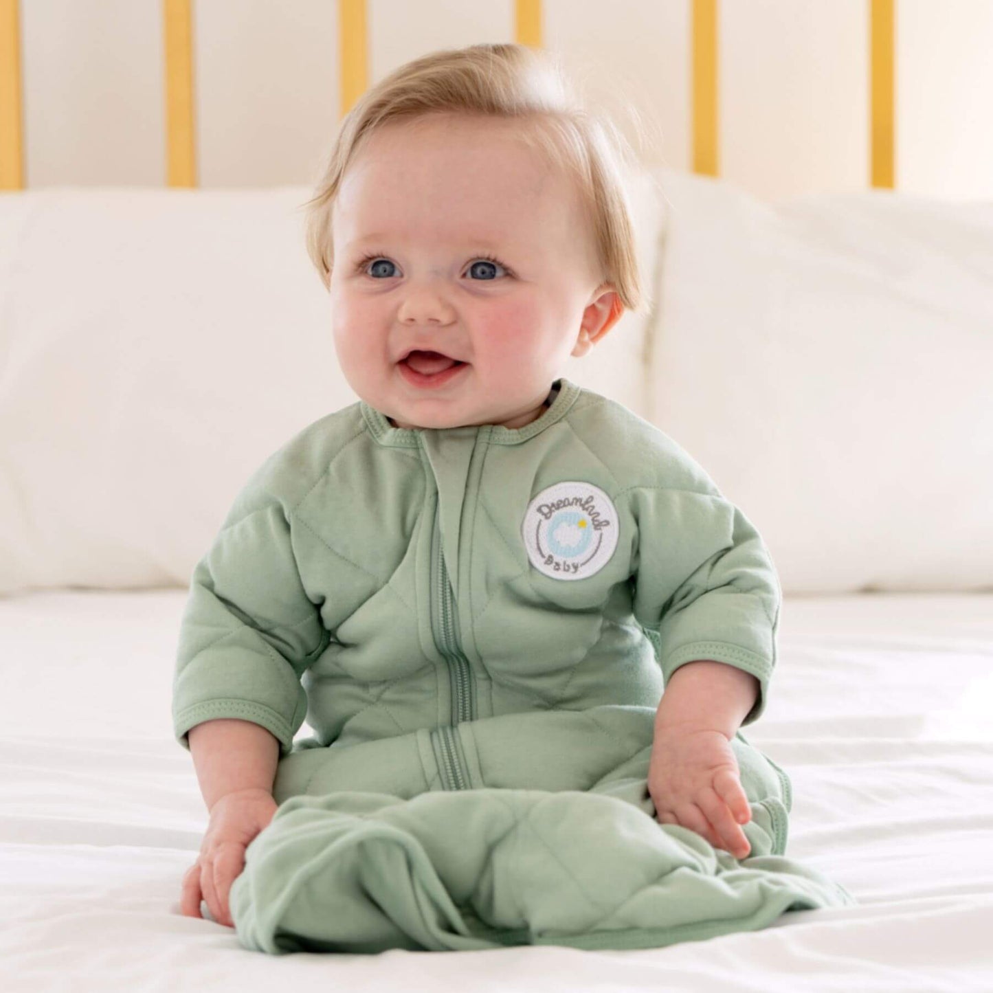 Baby wearing Dream Weighted Transition Swaddle - Sage Green, sitting on a white bed.