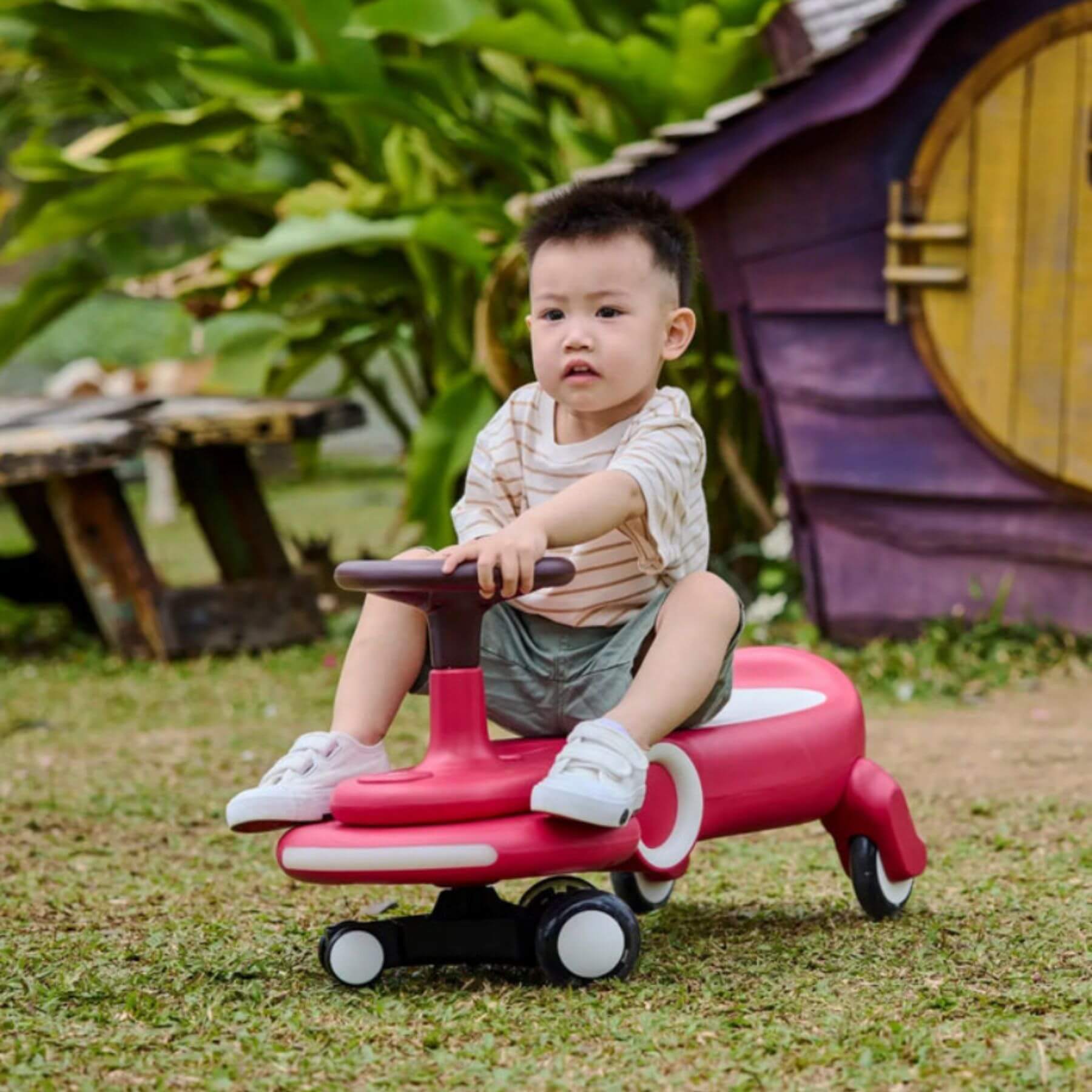 Child sitting on Hape Wiggle & Giggle Car – Red in an outdoor setting with greenery and a small wooden house.