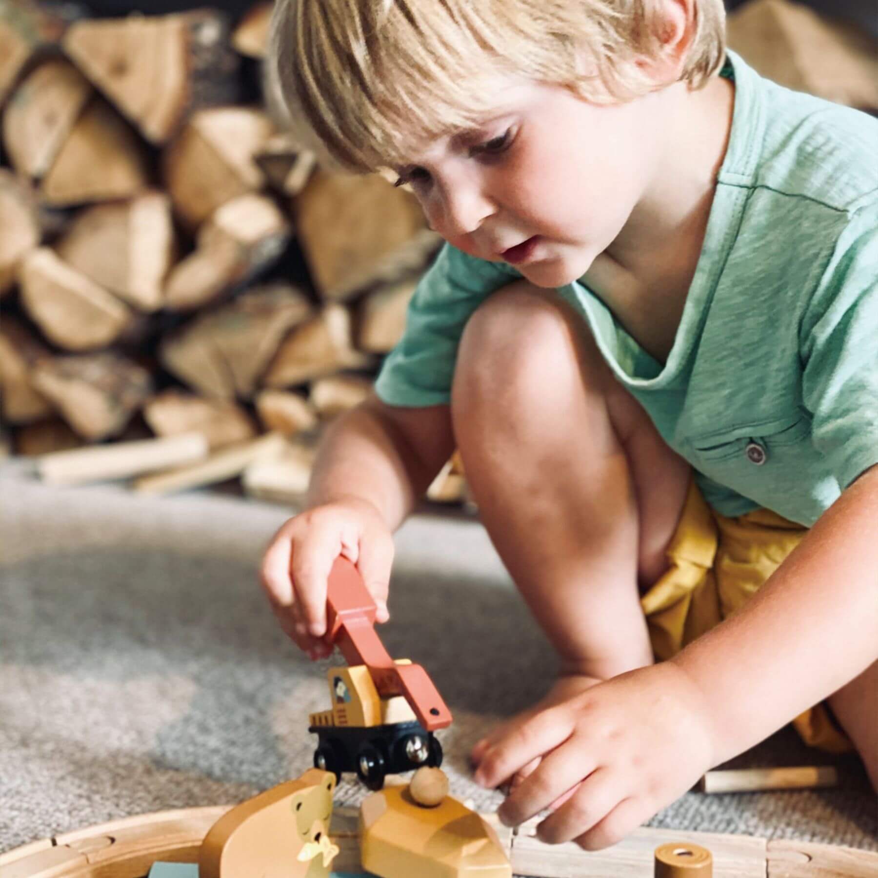 Child playing with a wooden toy train set on a carpeted floor with stacked firewood in the background.
