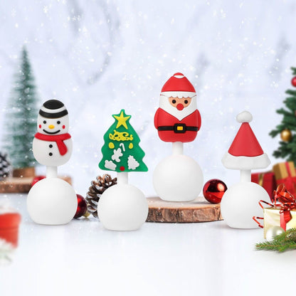 Four small white holiday-themed breast pump stoppers, including a snowman, Christmas tree, Santa Claus, and Santa hat, arranged on a snowy white surface with festive decorations in the background.