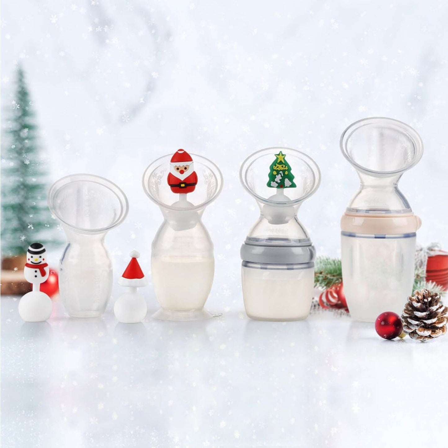 Four clear silicone breast pump flanges with different decorative holiday-themed nipple inserts, arranged on a snowy white surface with Christmas decorations in the background.