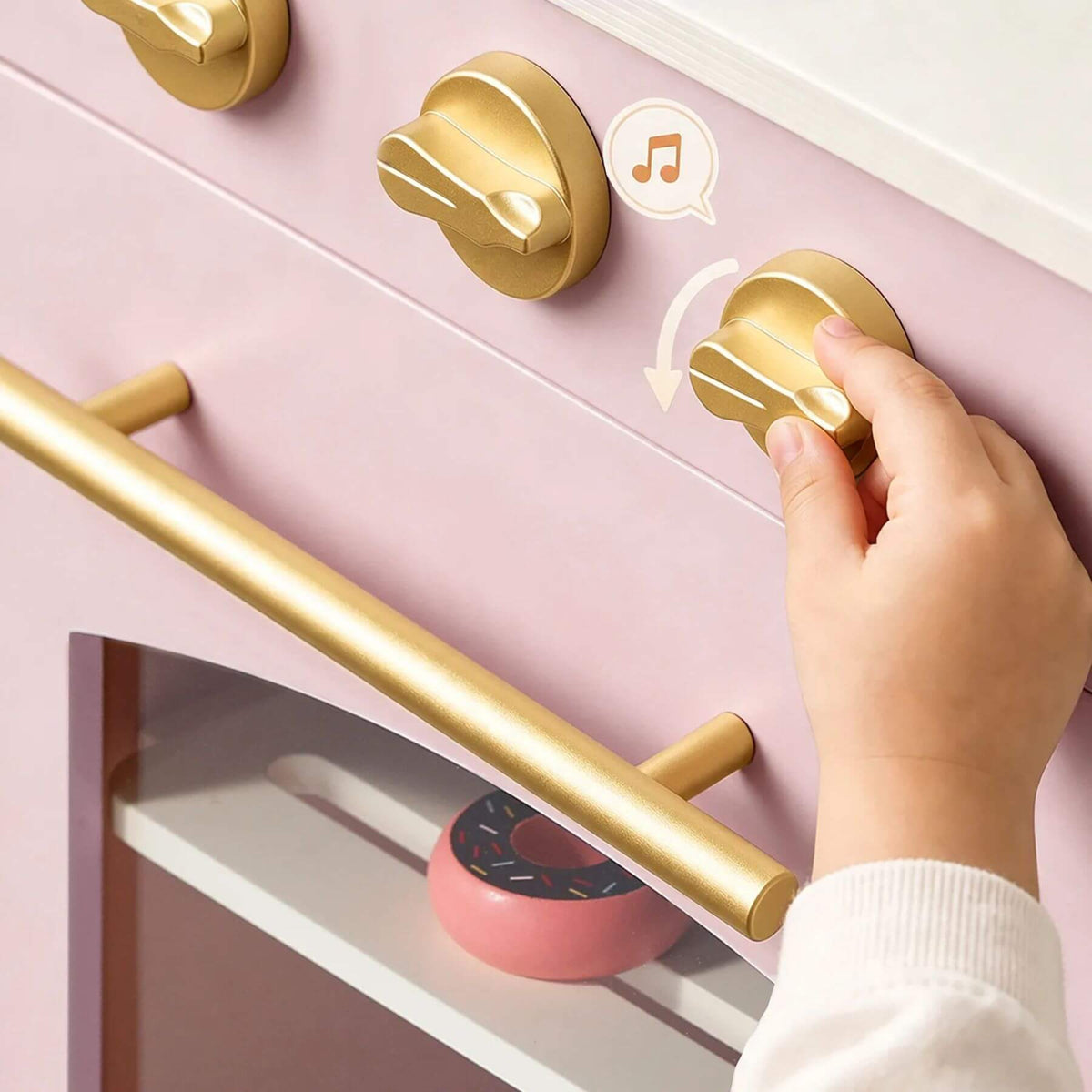 A child's hand turns a gold control knob on a pastel pink toy oven, with a small pink donut visible inside.
