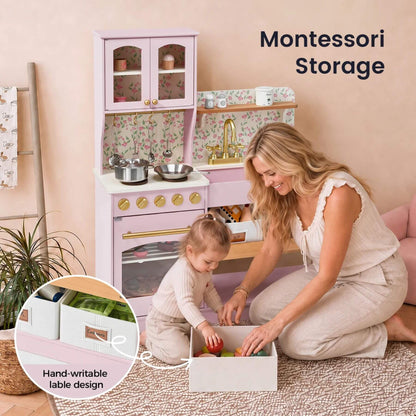 A woman and child are organizing toys in pastel pink Montessori storage boxes on a woven rug, with a matching pink play kitchen in the background.