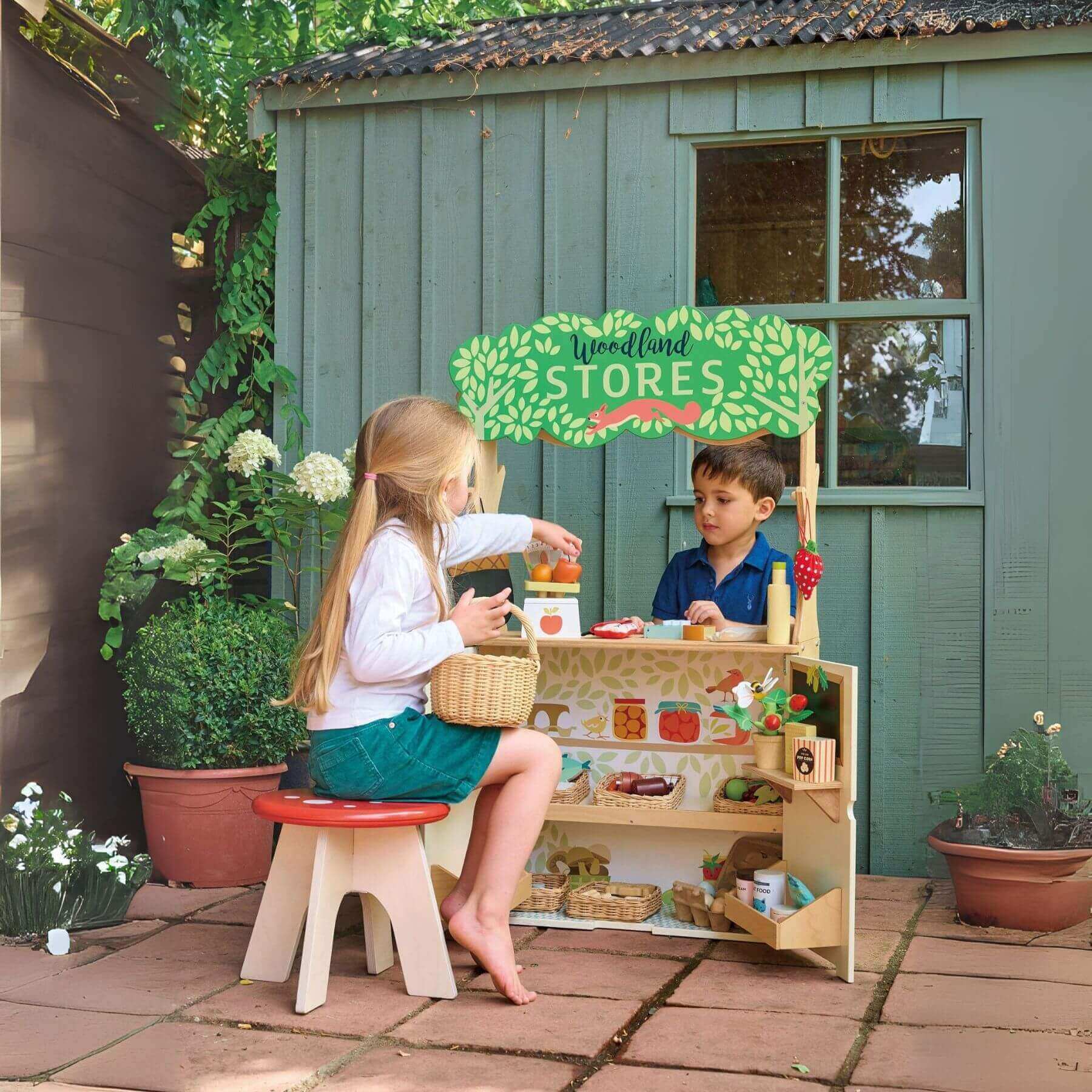 Two children playing with Tender Leaf Woodland Stores and Theater outdoors next to a green building.