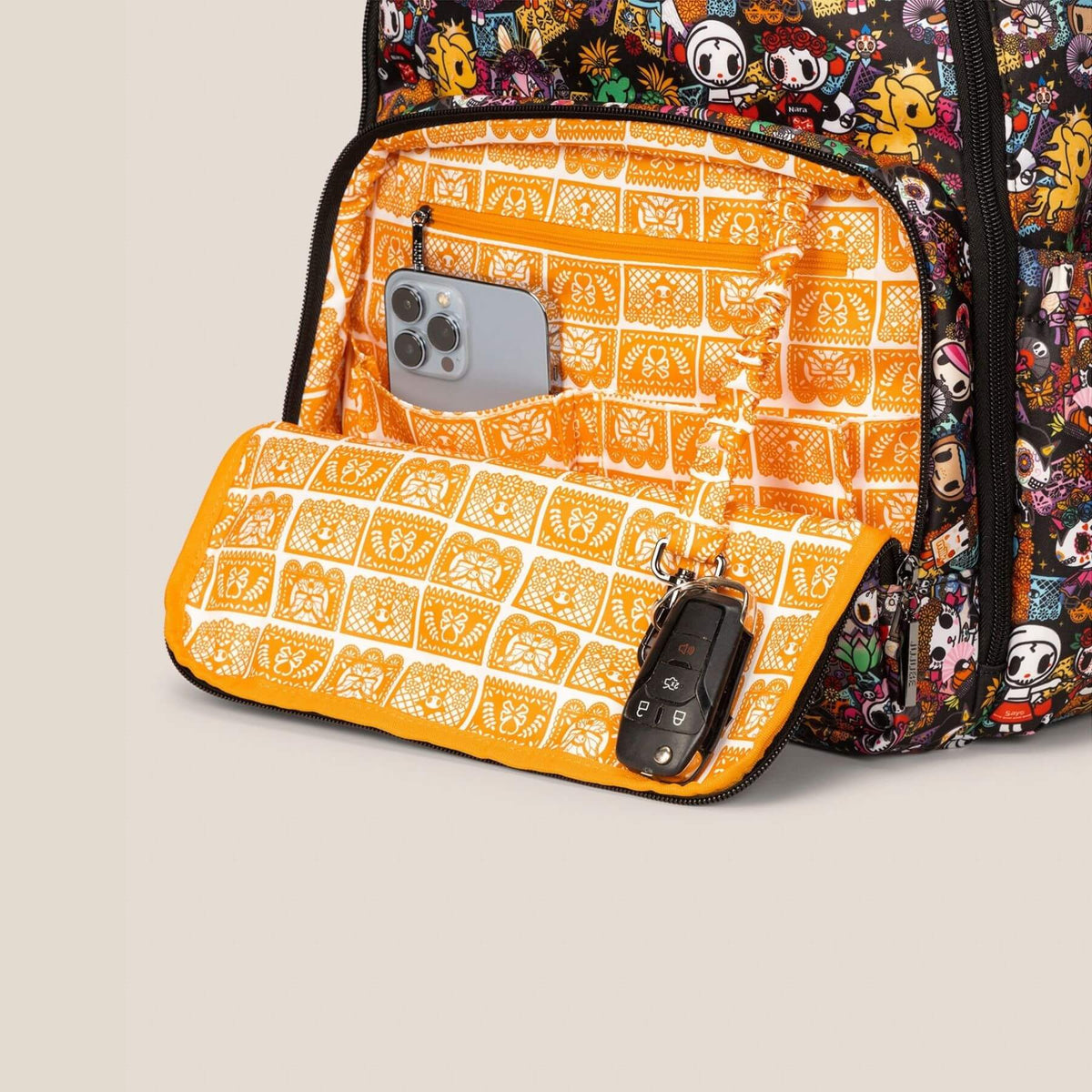 A close-up of tokidoki Zealous Backpack Diaper Bag – Dia de los Muertos pocket shown open, revealing an orange and white patterned interior organizer with a phone pocket and a key fob, against a light beige background.