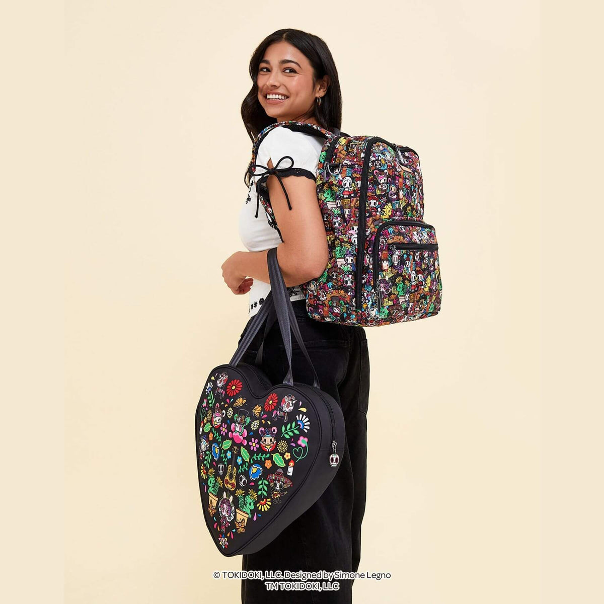 A woman wearing a white t-shirt and black pants is shown from the back, carrying tokidoki Zealous Backpack Diaper Bag – Dia de los Muertos and holding a matching heart-shaped tote bag.